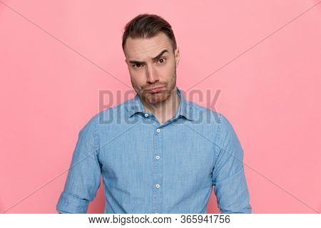 casual man looking at camera and raising one eyebrow, wearing a blue shirt and standing on pink studio background