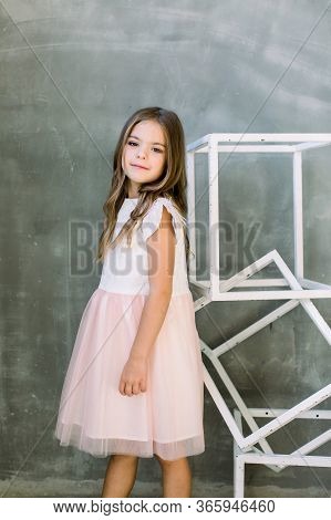 Beautiful Little Girl With Broun Long Hair On A Grey Background Near The White Constructure Of The C