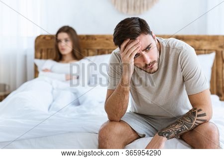 Problem In Bed. Man Sits On Bed And Holds On His Head, Woman Is Offended And Crossed Her Arms