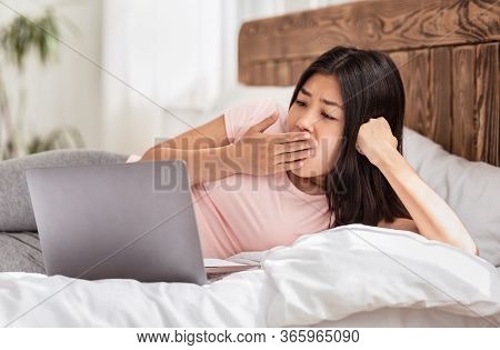 Bored Asian Girl Using Laptop Lying In Bed At Home In The Morning, Yawning And Browsing Internet. We