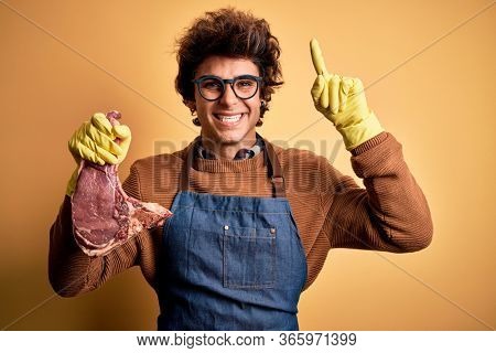 Young handsome butcher man holding meet steak standing over isolated yellow background smiling amazed and surprised and pointing up with fingers and raised arms.
