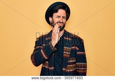 Young handsome hispanic bohemian man wearing hippie style and boho hat touching mouth with hand with painful expression because of toothache or dental illness on teeth. Dentist