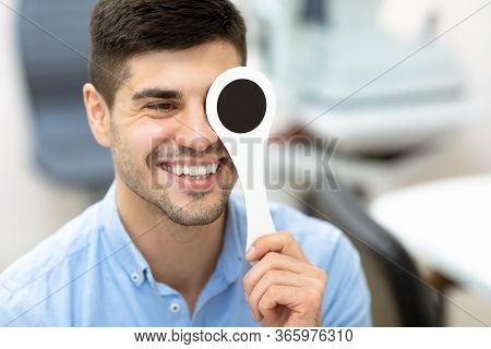 Cheerful Male Patient Doing Eye Test In Clinic, Holding Occluder And Looking At Chart, Copy Space