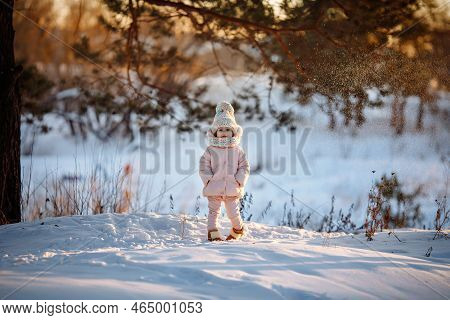 An Adorable Little Girl In A Warm Coat And A Colorful Hat With A Pom-pom Stands In The Snow On The S