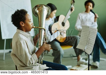 Side View Of Pre-teen Schoolboy Blowing Wind Instrument Against Two Intercultural Classmates Playing