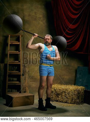Retro Circus. Cinematic Portrait Of Retro Circus Strongman Wearing Striped Sports Swimsuit Holding B