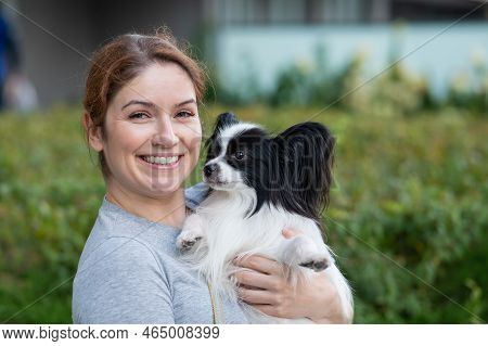 Caucasian Red-haired Woman Holding Pappilion Dog In Her Arms Outdoors. Black And White Continental S