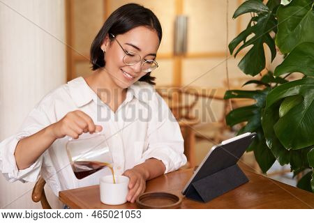 Freelance And Remote Workers. Smiling Young Woman Pouring Coffee In A Cup, Sitting In Cafe And Looki