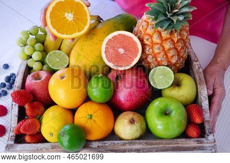 Wooden Basket On White Table Full Of Fresh Colorful Fruits. Mature Woman Holding Half Orange Fruit. 
