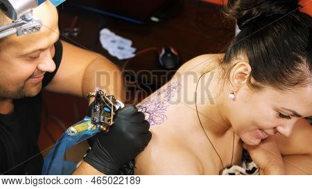 In A Tattoo Salon , A Specialist Is Doing A Tattoo On Womans Back, A Floral Ornament. A Man Works In