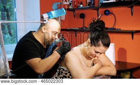 In A Tattoo Salon , A Specialist Is Doing A Tattoo On Womans Back, A Floral Ornament. A Man Works In