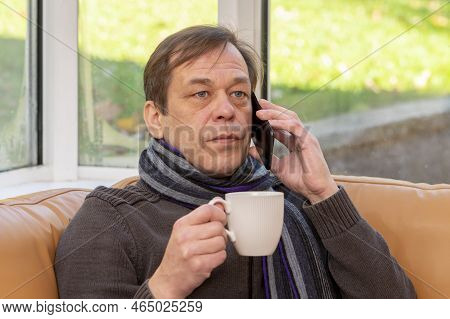Portrait Of A Serious Man 45-50 Years Old With A White Mug In His Hand And A Scarf, Talking On A Sma