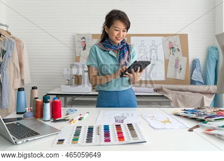 Young Asian Dressmaker In Striped Scarf Checking Email Order From Tablet Computer. Laptop Computer, 