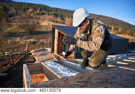 Male Builder Doing Thermal Insulation On Roof Of Wooden Frame House. Man Worker Spraying Polyurethan