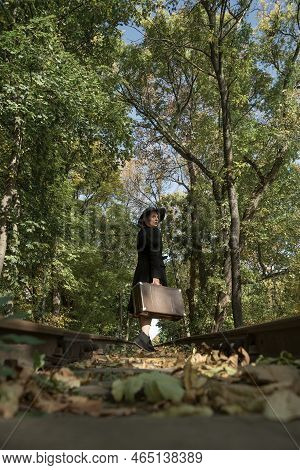Stylish Woman Walking On Rails, Walking Through Forest. Broad-brim Hat, Black Coat And Retro Valise 