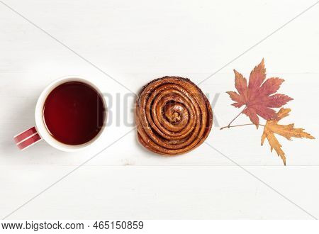 A Cup With A Hot Drink, A Cinnamon Bun And Autumn Leaves On A Light Wooden Background. Close-up