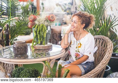 Young Woman With Blond Curly Hair Sitting On A Wicker Chair With Many Plants Admires The Vegetation.