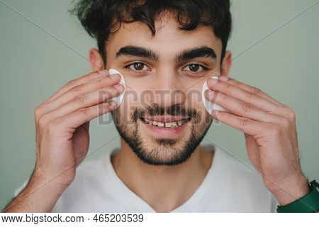 Handsome Man Cleaning Face Skin With Cotton Pads Over Green Background And Looking At Camera. Lookin