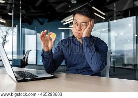 Upset And Dissatisfied Young Man Asian Businessman Holds A Bitcoin In His Hands, Looks At It Sadly. 