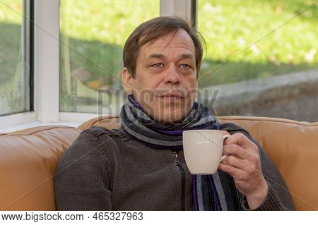 An Elderly Man Of 45-50 Years Old In A Scarf Holds A White Mug In His Hand While Sitting On The Sofa