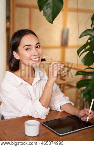 Beautiful Asian Girl Sitting In Cafe With Cup Of Coffee, Drawing On Digital Tablet With Graphic Pen,