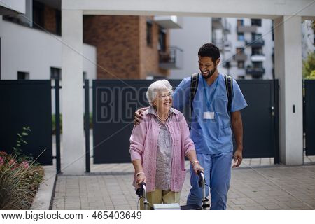 Caregiver Walking With Senior Woman Client In Front Of Nurishing Home.