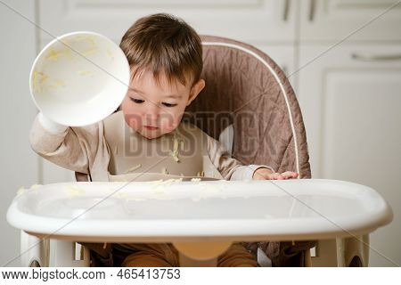 A Funny Child Is Eating Sitting On A Kitchen Chair Picked Up The Plate To Throw. Hungry Baby Boy Eat
