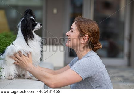 Caucasian Red-haired Woman Cuddling With Pappilion Dog Outdoors. Black And White Continental Spaniel