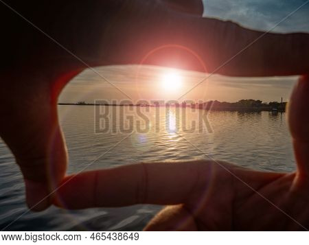 Gesture Formed By Fingers Over The Setting Sun On The Sea. Lensflare From The Low Sun Above Calm Sea