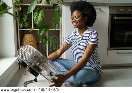 Happy Smiling Young Black Woman Enjoying Fresh Air While Cooling By Electric Fan At Home, Sitting Ne