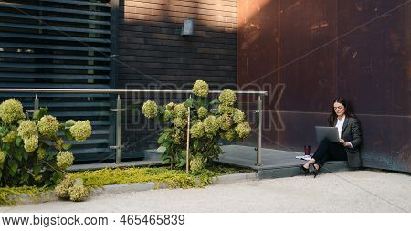 Far Away View Of A Young Woman Sitting Outdoor On Terrace, Working Remotely Online On Laptop. Freela