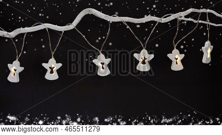 Glowing Garland In The Form Of White Wooden Angels On A Tree Branch On A Black Background. Snow Bord