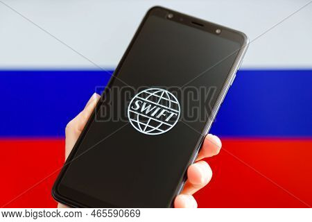 Ukraine, Kiev - February, 23 2022: Hand Holding Mobile With Swift Logo And Russian Flag In Backgroun