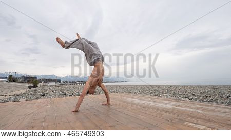 Shirtless Caucasian Man Doing Backflip On Pebble Beach.