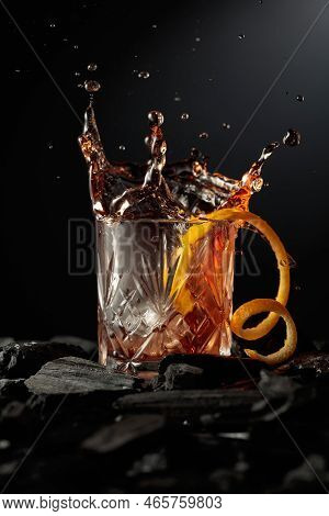Piece Of Ice Falls Into The Glass With A Cocktail. Cocktail With Ice And Orange Peel On A Dark Backg