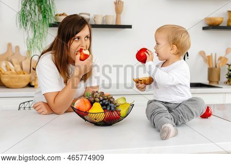 Mom And Her Son Are In The Kitchen Eating Healthy Food. The Baby Is Holding A Cookie And An Apple. R