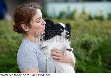 Caucasian Red-haired Woman Holding Pappilion Dog In Her Arms Outdoors. Black And White Continental S