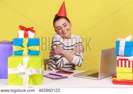 Satisfied Woman Office Employee With Party Cone Sitting At Workplace Surrounded By Many Presents And
