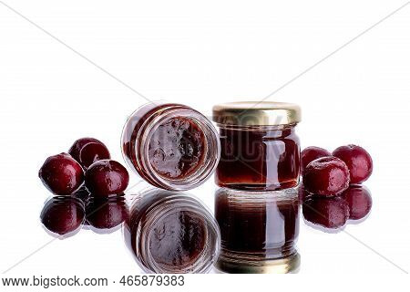 Cherry Jam In A Jar On A White Mirror Background With Cherry Fruits And Their Reflection. Concept Of