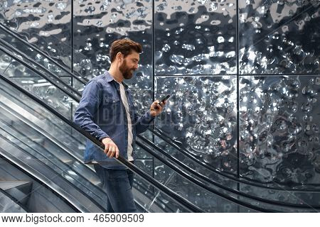 Smiling Handsome Man Using Smartphone, While Going Down On Escalator Of Shopping Mall. Side View Of 