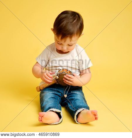 Toddler Baby Plays A Hand Drum, A Child With A Percussion Musical Instrument On A Studio Yellow Back