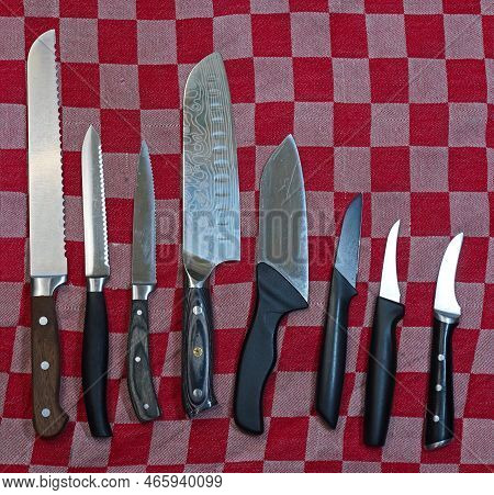 Eight Kitchen Knives On A Red-white Checkered Towel. The First Is A Bread Knife. Next A Tomato Knife