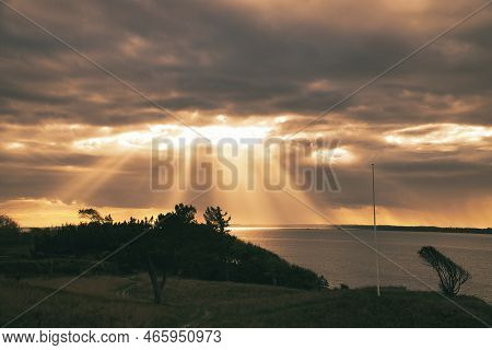 On The Coast Of Hundested. Sun Rays Break Through The Dramatic Sky Through The Clouds. Meadow With P
