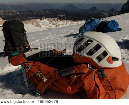 Several Backpacks And A Protective Helmet Lie On A Snowy Slope, Austria, Alps, Dachstein