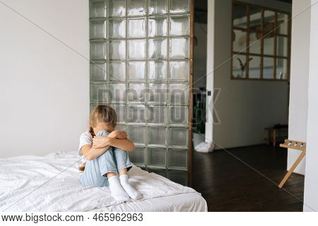 Sad Unrecognizable Little Girl Hugging Knee, Sobbing With Head Bowed And Crying Sitting On Bed In Be