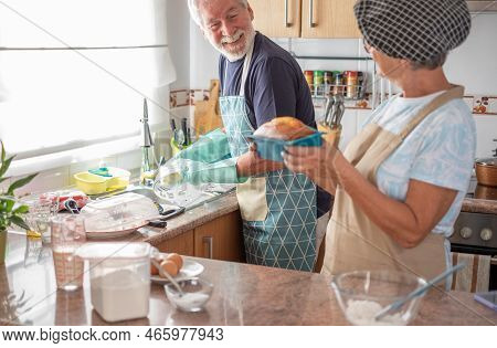 Cheerful Senior Couple In The Home Kitchen While Wife Holds A Freshly Baked Homemade Plumcake And Hu