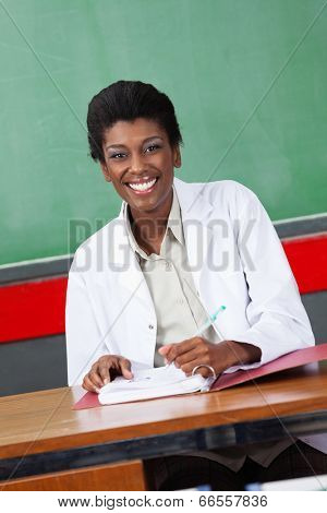 Portrait of happy African American female teacher with pen and binder sitting at desk in classroom