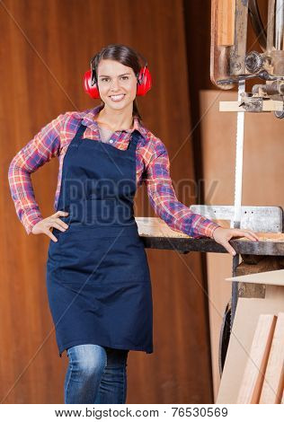 Portrait of confident female carpenter with hand on waist standing by bandsaw in workshop