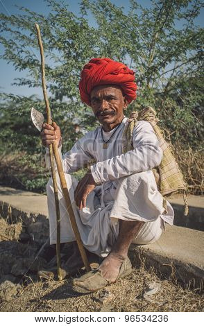 GODWAR REGION, INDIA - 14 FEBRUARY 2015: Elderly Rabari tribesman with red turban sits and holds ax and stick. Post-processed with grain, texture and colour effect.