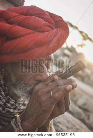 GODWAR REGION, INDIA - 14 FEBRUARY 2015: Elderly Rabari tribesman with red turban and blanket smokes chillum. Post-processed with grain, texture and colour effect.
